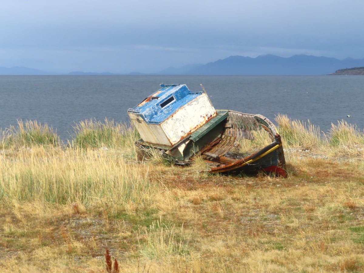 Weathered boat on grassy shore with ocean and mountains in background Punta Arenas Chile