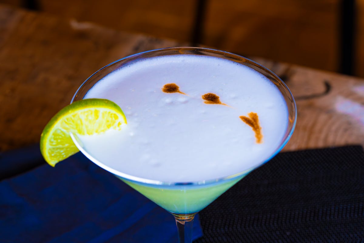 Close-up of a pisco sour cocktail with lime garnish and angostura bitters drops on a rustic wooden bar