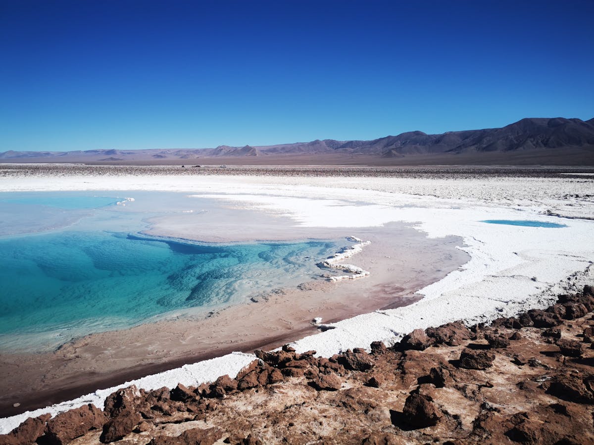 Turquoise lagoon surrounded by red and brown desert terrain with snow-capped mountains in the Atacama