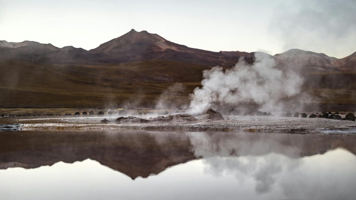 El Tatio geyser field with steam columns reflected in calm water and mountains behind